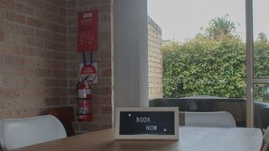 A room with a brick wall, a fire extinguisher, a window with a view of greenery, and a table with a sign that says "BOOK NOW". - A short walk to Black Head Beach, park with bbqs & amenities, shops and cafes (Black Head)