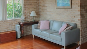 A living room with a brick wall, a ceiling fan, a window with curtains, a sofa, and a lamp. - A short walk to Black Head Beach, park with bbqs & amenities, shops and cafes (Black Head)