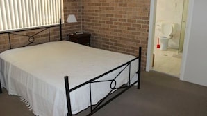 A bedroom with a metal bed frame, a bedside table, a lamp, and a brick wall. - A short walk to Black Head Beach, park with bbqs & amenities, shops and cafes (Black Head)