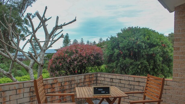An outdoor seating area with a table and two chairs, a brick wall, and a sign that reads "BOOK NOW". - A short walk to Black Head Beach, park with bbqs & amenities, shops and cafes (Black Head)