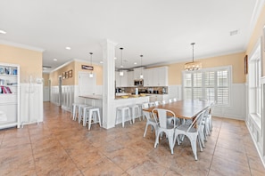 A spacious kitchen with white cabinetry, a dining area with a wooden table and white chairs, and a large window with shutters. - Landshark, Luxury Oceanfront Cherry Grove Home with Game Room and Pool (North Myrtle Beach)