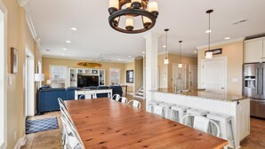 A spacious kitchen with a wooden dining table, white bar stools, a stainless steel refrigerator, and a large island with a sink. - Landshark, Luxury Oceanfront Cherry Grove Home with Game Room and Pool (North Myrtle Beach)