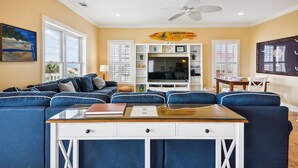 A living room with a large blue sofa set, a wooden coffee table, and a flat-screen TV. There is a ceiling fan, a window with blinds, and a sign above the TV. - Landshark, Luxury Oceanfront Cherry Grove Home with Game Room and Pool (North Myrtle Beach)