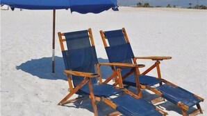 Zwei Holzliegen mit blauen Kissen und einem blauen Sonnenschirm an einem Sandstrand.