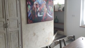 A dining area with a wooden table and chairs, a colorful elephant painting on the wall, and a white door with a textured finish. - house for 4 to 6 persons fenced garden (Sainte-Jalle)