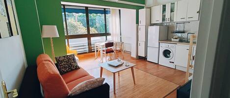 A compact living space with a sofa, dining table, chairs, a washing machine, and a kitchen area.