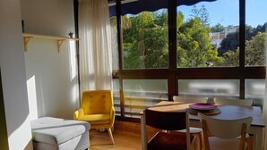 Una habitación acogedora con sofá, un sillón amarillo y una mesa de comedor con sillas. Amplias ventanas ofrecen vistas al exterior con vegetación.
