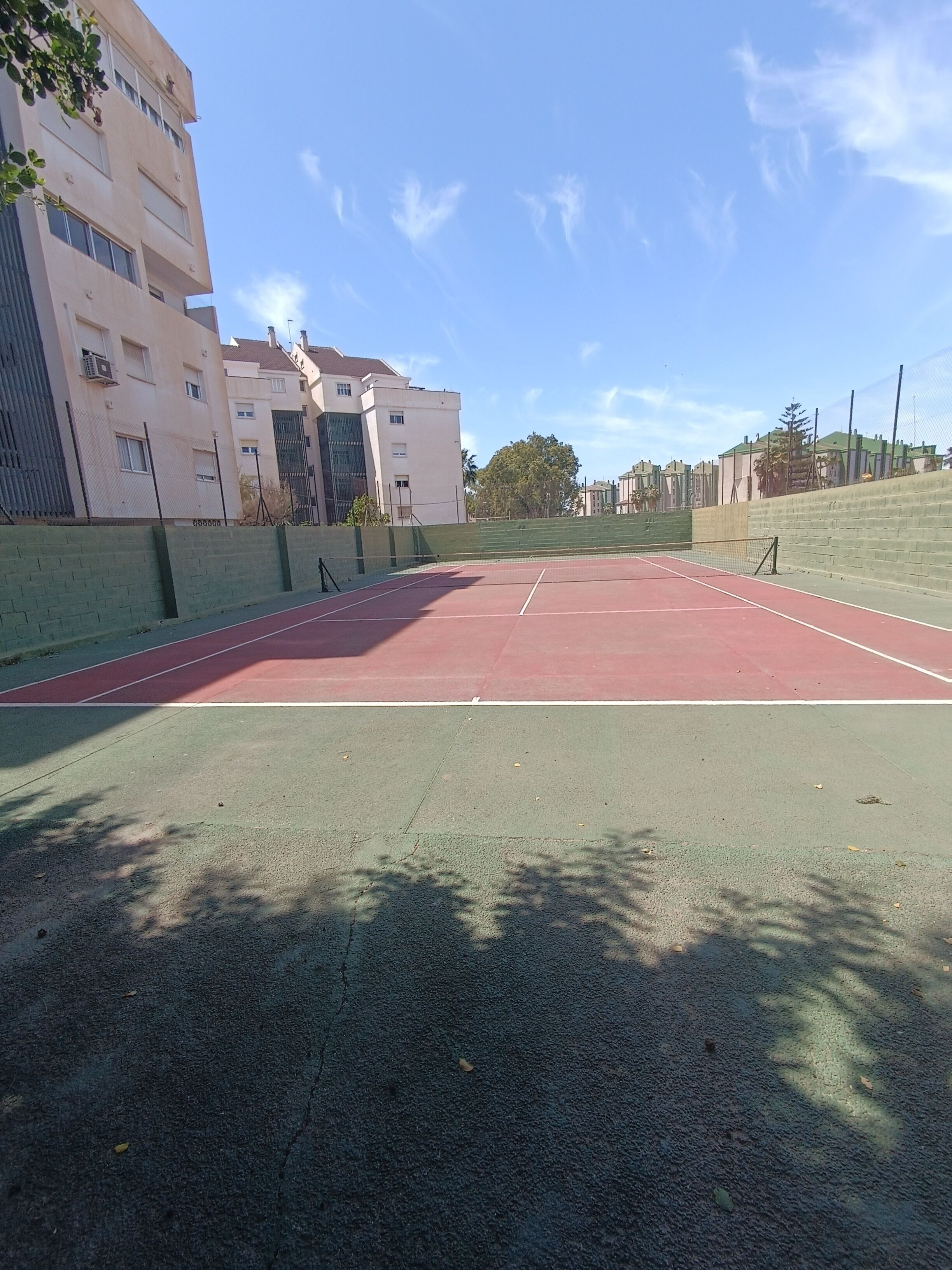 An outdoor tennis court with a green and red surface, surrounded by a low brick wall and a fence.