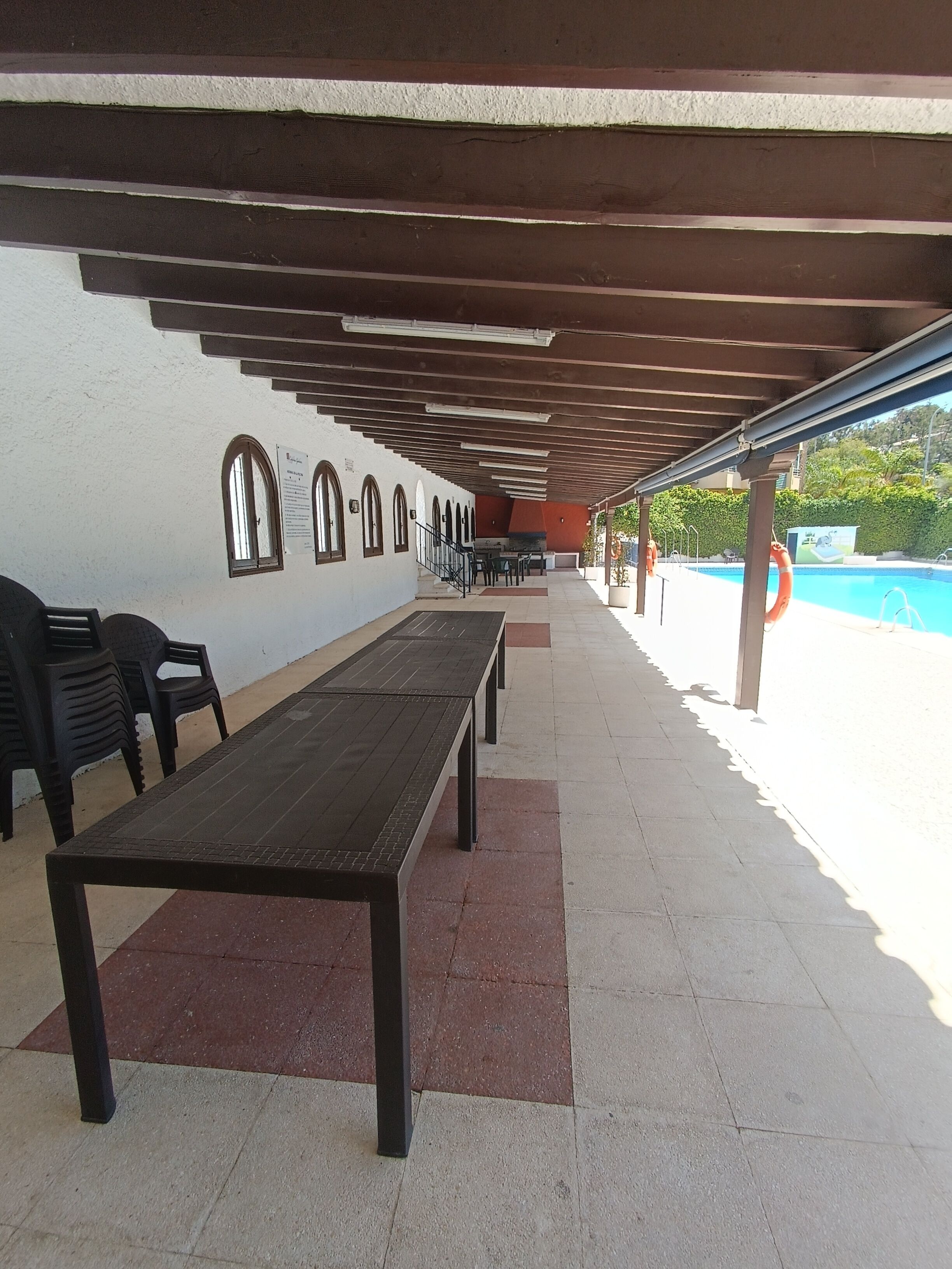 An outdoor covered patio area with long tables and chairs, a pool in the background, and a white wall with arched windows.