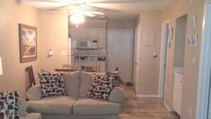 A living room with a beige sofa, patterned pillows, a tufted ottoman, a ceiling fan, and a kitchen in the background. - 1st Floor, Updated, 5 min walk to the beach! (Hilton Head Island)