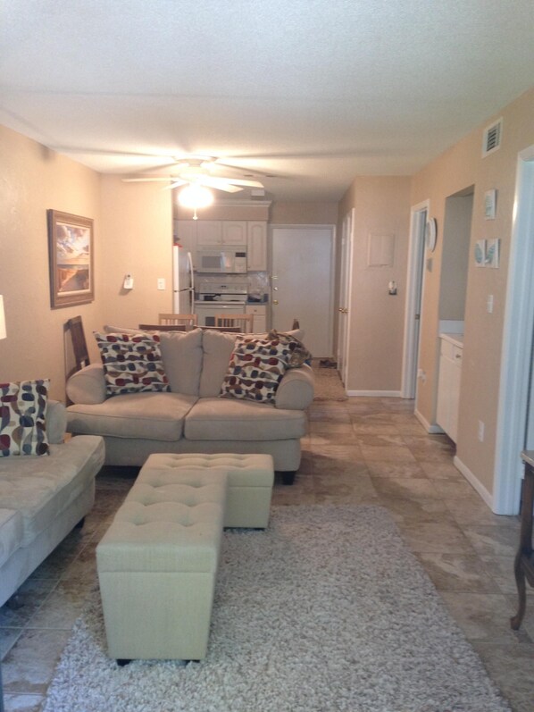 A living room with a beige sofa, patterned pillows, a tufted ottoman, a ceiling fan, and a kitchen in the background.