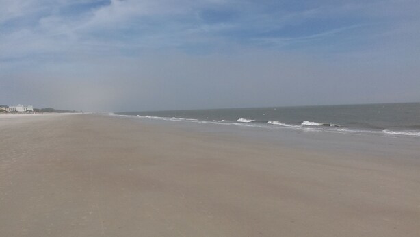 A wide sandy beach with gentle waves, a clear sky, and buildings in the distance. - 1st Floor, Updated, 5 min walk to the beach! (Hilton Head Island)