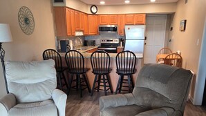 A modern kitchen with wooden cabinets, a microwave, and a refrigerator. There are four black chairs around a central island. - Oceanfront Cherry Grove 2 BR Condo - Excellent Reviews!!  (North Myrtle Beach)