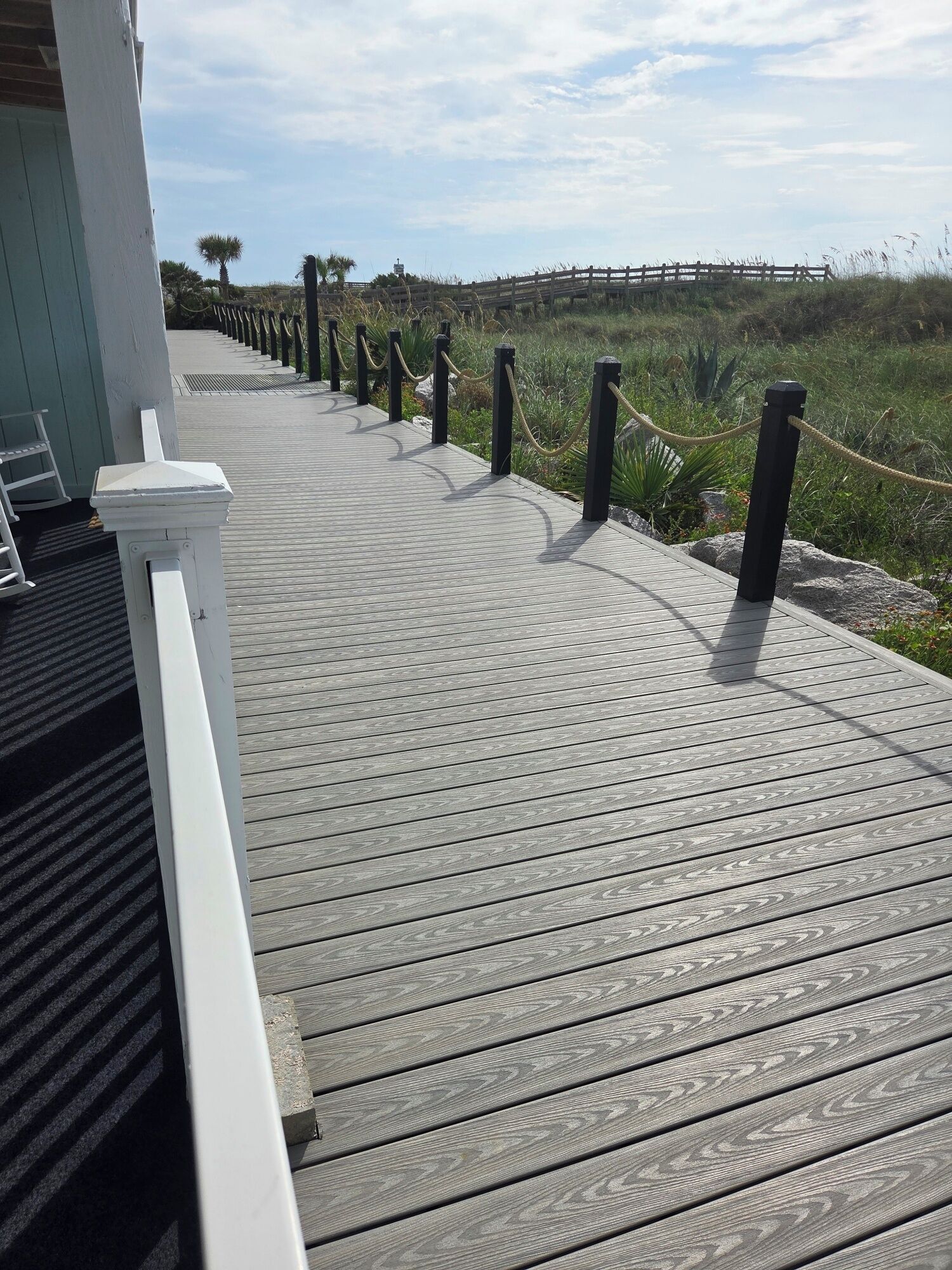 A wooden boardwalk with black bollards and a rope barrier, leading to a grassy area with a fence in the background.