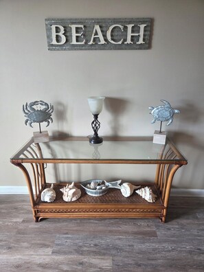 A glass-top table with a rustic wooden base, decorated with seashells and a crab figurine, against a wall with a 'BEACH' sign. - Oceanfront Cherry Grove 2 BR Condo - Excellent Reviews!!  (North Myrtle Beach)