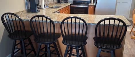 A kitchen with wooden cabinets, a stainless steel refrigerator, a microwave, a stove, and a sink. There are four black chairs at the island.