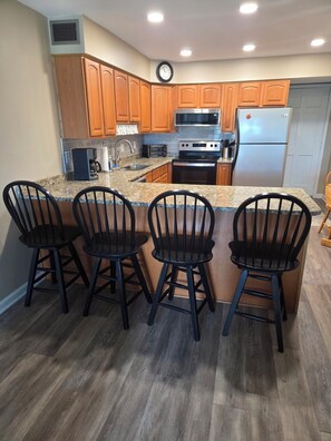 A kitchen with wooden cabinets, a stainless steel refrigerator, a microwave, a stove, and a sink. There are four black chairs at the island. - Oceanfront Cherry Grove 2 BR Condo - Excellent Reviews!!  (North Myrtle Beach)