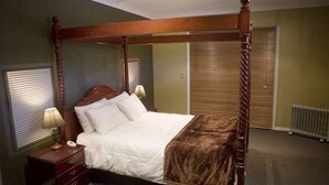 A four-poster bed with a wooden frame, a white bedspread, and a brown throw blanket. There is a nightstand with a lamp and a telephone. The room has a radiator and a window with blinds. - Stanthorpe Country Resort, 6 bedrooms for 12 guests (Stanthorpe)