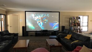 A living room with a large screen displaying 'AVATAR', a black leather sofa, a red sofa, a coffee table, and a bookshelf. - Stanthorpe Country Resort, 6 bedrooms for 12 guests (Stanthorpe)