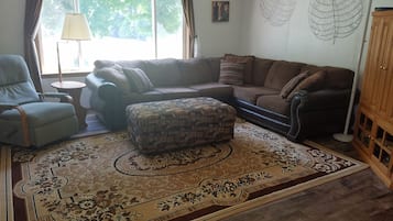 A living room with a sofa, ottoman, and armchair, a patterned rug, a lamp, and a window with curtains.