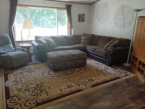 Una sala de estar con sofá, reposapiés y sillón, alfombra estampada, lámpara y ventana con cortinas.