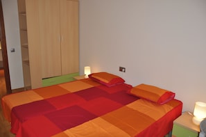 A bed with a red and orange quilt, a green nightstand with a lamp, and a wooden wardrobe.