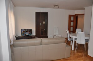 Ein Wohnzimmer mit einem beigefarbenen Sofa, einem Holzschrank, einem Esstisch mit Stühlen und einem Fernseher auf einem Schrank.