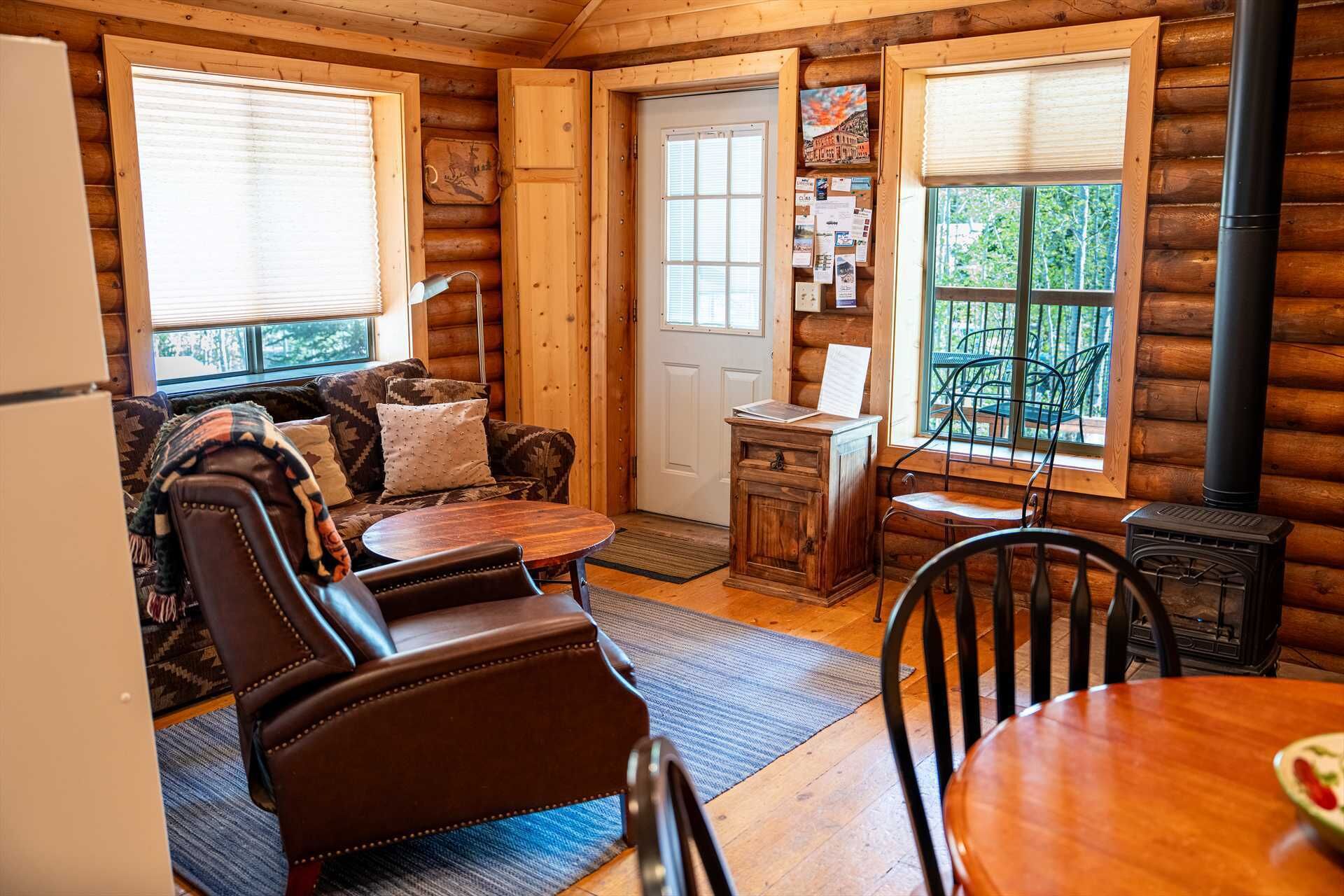L’intérieur chaleureux d’un chalet avec un mur en rondins de bois, un coin repas avec une table et des chaises, une cheminée et une fenêtre donnant sur la forêt.