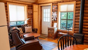 Un interior de cabaña acogedor con una pared de madera rústica, una zona de comedor con mesa y sillas, una chimenea y una ventana con vistas a árboles.