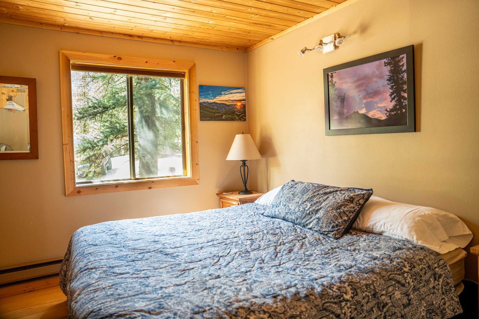 Une chambre avec un grand lit, une fenêtre donnant sur des arbres, une table de chevet avec une lampe et deux tableaux encadrés au mur.