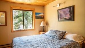 Un dormitorio con una cama grande, una ventana con vista a árboles, una mesita de noche con lámpara y dos cuadros enmarcados en la pared.