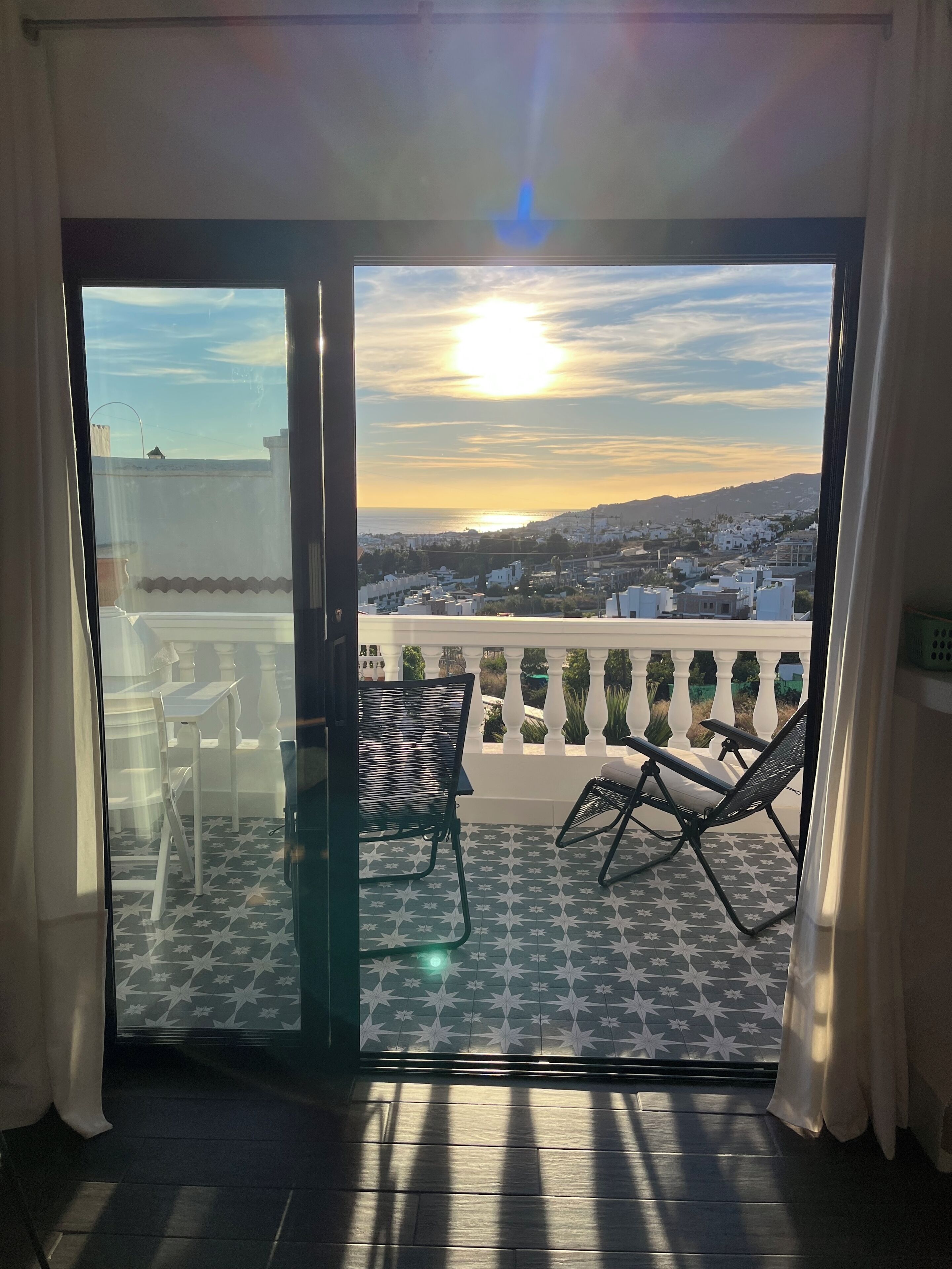 A balcony with a star-patterned floor, two chairs, and a view of the sunset over a cityscape.