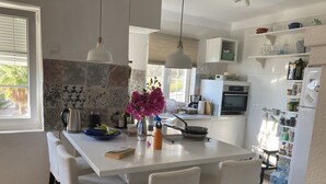 Une cuisine moderne avec des meubles blancs, un îlot central doté de chaises, un éclairage suspendu et une vue sur l’extérieur.