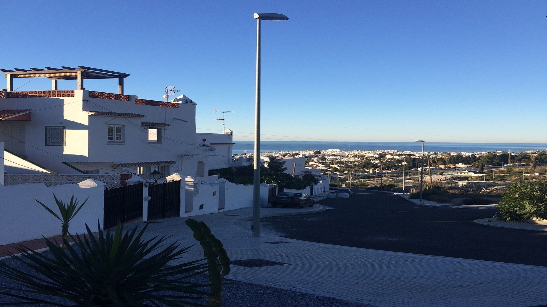 Un carrer residencial amb una vista despejada del mar i un paisatge urbà a la distància.