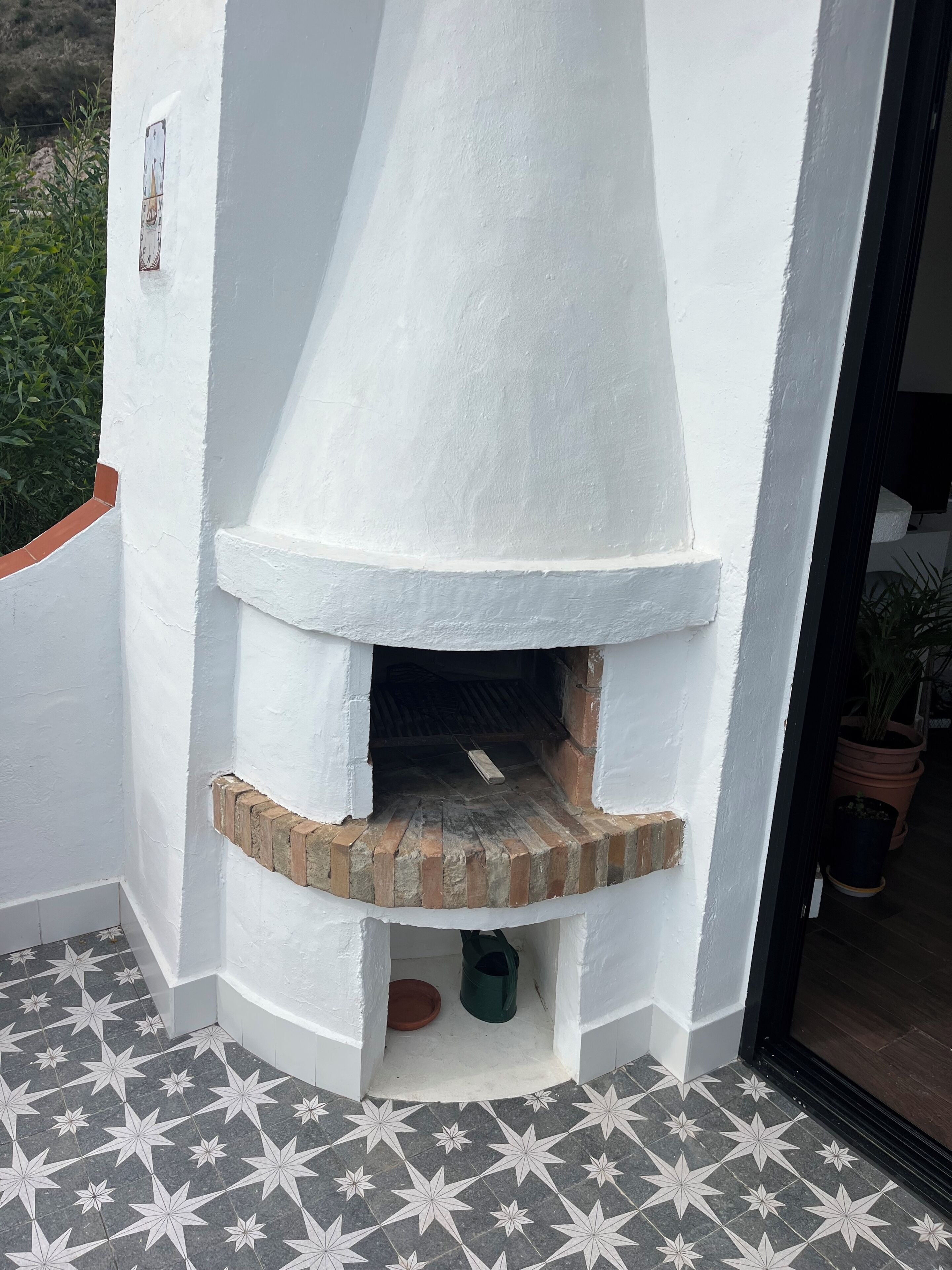 A white outdoor fireplace with a brick base, surrounded by a white wall and a tiled floor.
