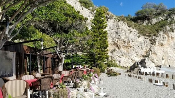 Un espace extérieur aménagé avec des chaises et des tables en osier, entouré de verdure et bordé d’une colline rocheuse.
