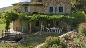 A stone house with green shutters, a covered patio with chairs, and a garden with various plants and flowers.