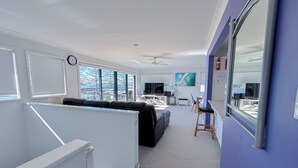A modern living room with a sofa, a dining area with chairs, a TV, and a large window with blinds. - Pet Friendly, Magnificent Ocean Views, A minutes' Easy Stroll To The Beach (Black Head)