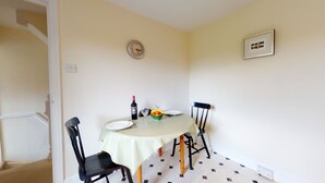 Un coin repas avec une table dressée pour deux personnes, agrémentée d’une nappe à carreaux, de deux chaises, d’une horloge, d’un tableau encadré et d’un saladier rempli de fruits.