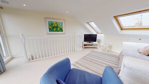 Une chambre avec un plafond en pente, une balustrade blanche, un téléviseur, une petite table et un lit agrémenté d’un coussin à motifs.