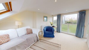 Un salon spacieux avec un canapé blanc, un fauteuil bleu et une vue sur un jardin verdoyant, grâce à de grandes fenêtres.