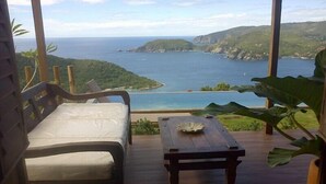 A room with a wooden floor, a white sofa, and a wooden table with a decorative item. A large window offers a view of a coastal landscape with cliffs and the sea.