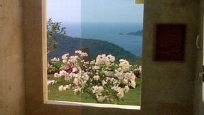 A room with a window overlooking a garden with pink flowers and a distant view of a coastline.