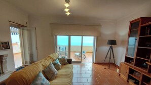 Un salon avec un canapé, une lampe et une bibliothèque. Une porte-fenêtre coulissante donne sur un balcon d’où l’on peut admirer la mer.