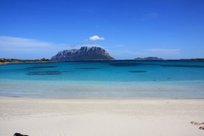 A beach with clear turquoise water, white sand, and a prominent rocky island in the background.