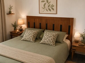 A neatly made bed with two pillows, a bedside lamp, a small table with a vase and candle, and a framed botanical print on the wall.