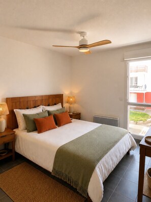 A bedroom with a bed, a ceiling fan, a nightstand with a lamp, and a window with a view of greenery outside.