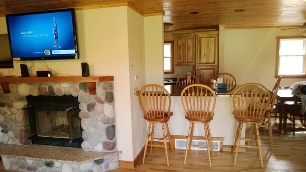A living room with a stone fireplace, wooden chairs, a television, and a kitchen area with wooden cabinets and a window.