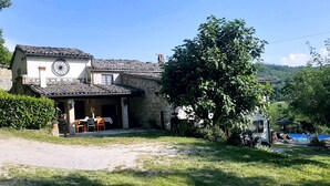 Un bâtiment rustique en pierre, doté d’un toit en tuiles, d’un espace repas avec des sièges en plein air et d’un grand arbre au premier plan.