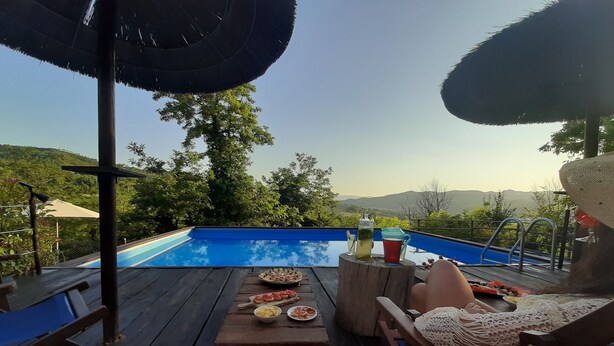 Une terrasse en bois avec une piscine, une table à manger et des chaises, offrant une vue panoramique sur un paysage pittoresque, avec des arbres et des collines.